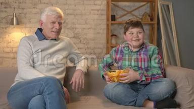 爷爷和孙子坐在沙发上看电视，胖男孩给爷爷吃薯片